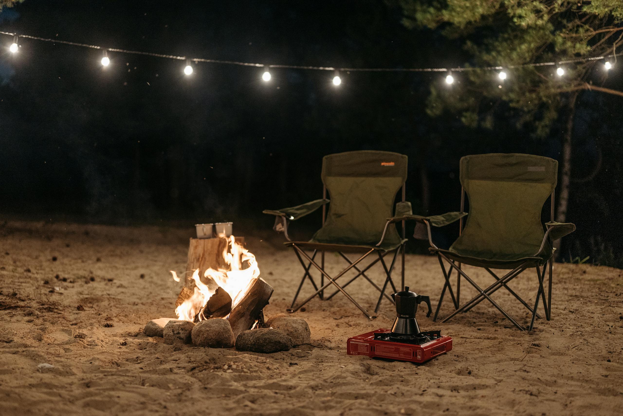 Outdoor camping scene with chairs and bonfire under string lights at night.